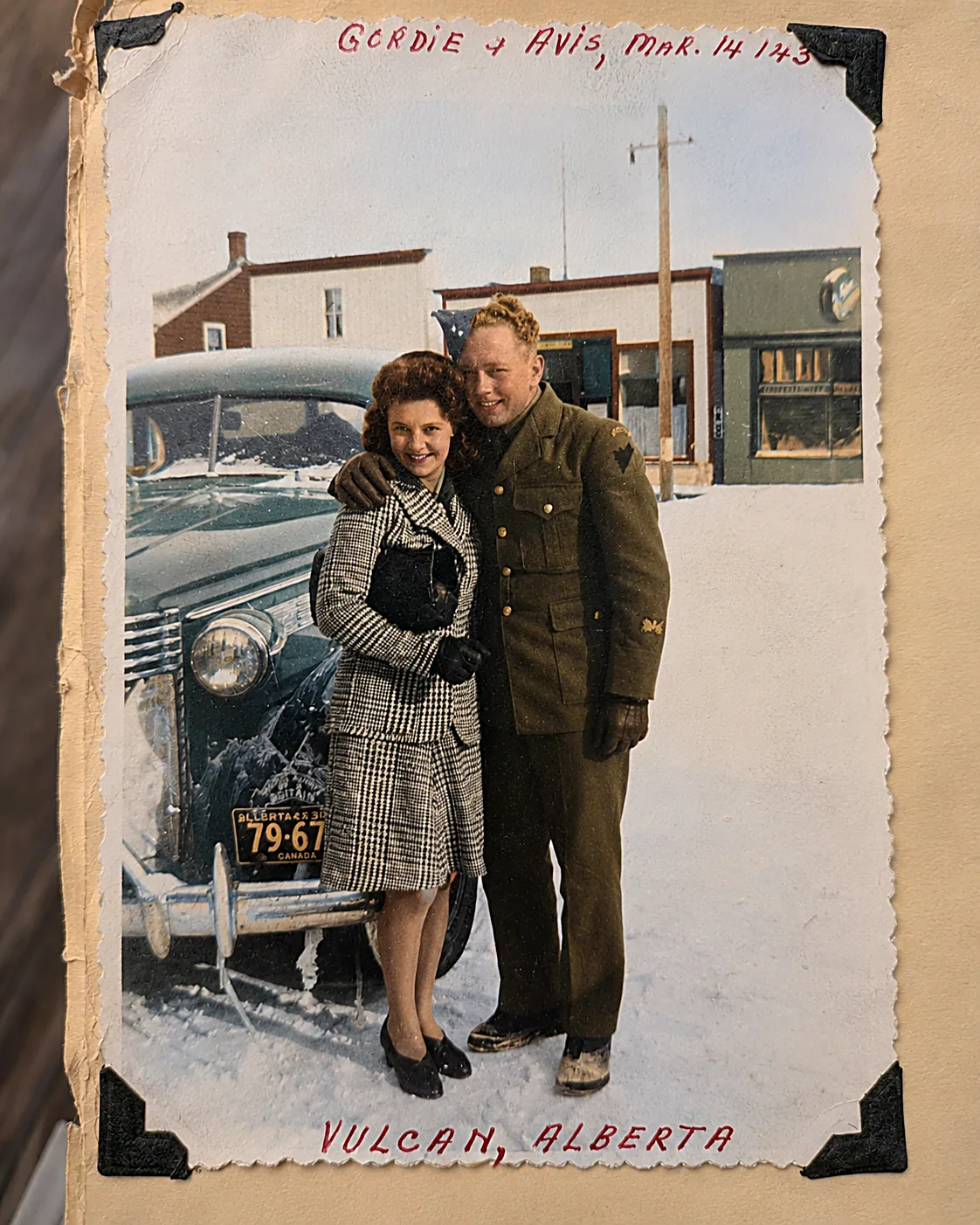 Colourised photograph of Gordie in his army uniform and Avis in a checked coat, arm in arm in the snow beside a green car on a small-town street. Caption in the album reads Gordie & Avis, Mar. 14/43, Vulcan, Alberta.