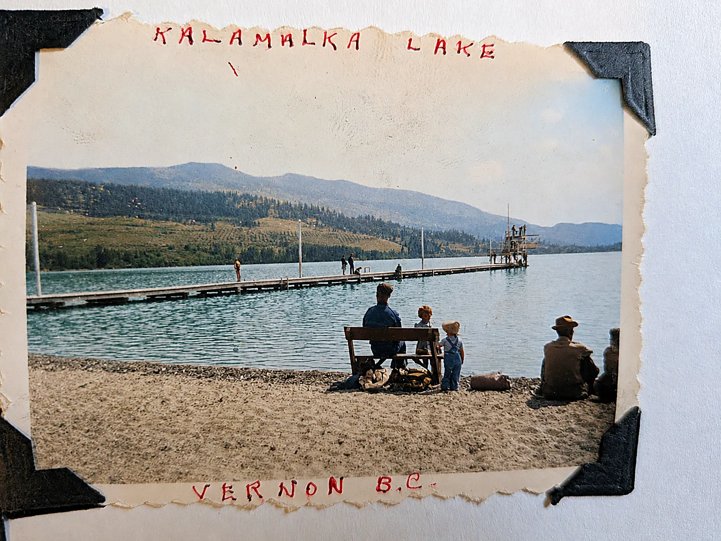 Colourised photograph looking across Kalamalka Lake to green hills, a long wooden dock and diving platform, with a man and two small children on the beach in the foreground.
