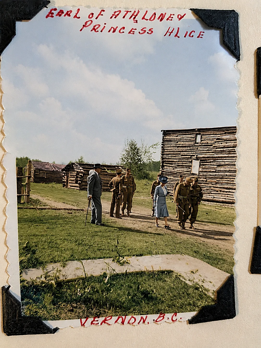 Colourised photograph of a man in a suit with a cane and a woman in a blue coat and hat walking an inspection past soldiers and log huts at an army camp. The album labels them the Earl of Athlone and Princess Alice.