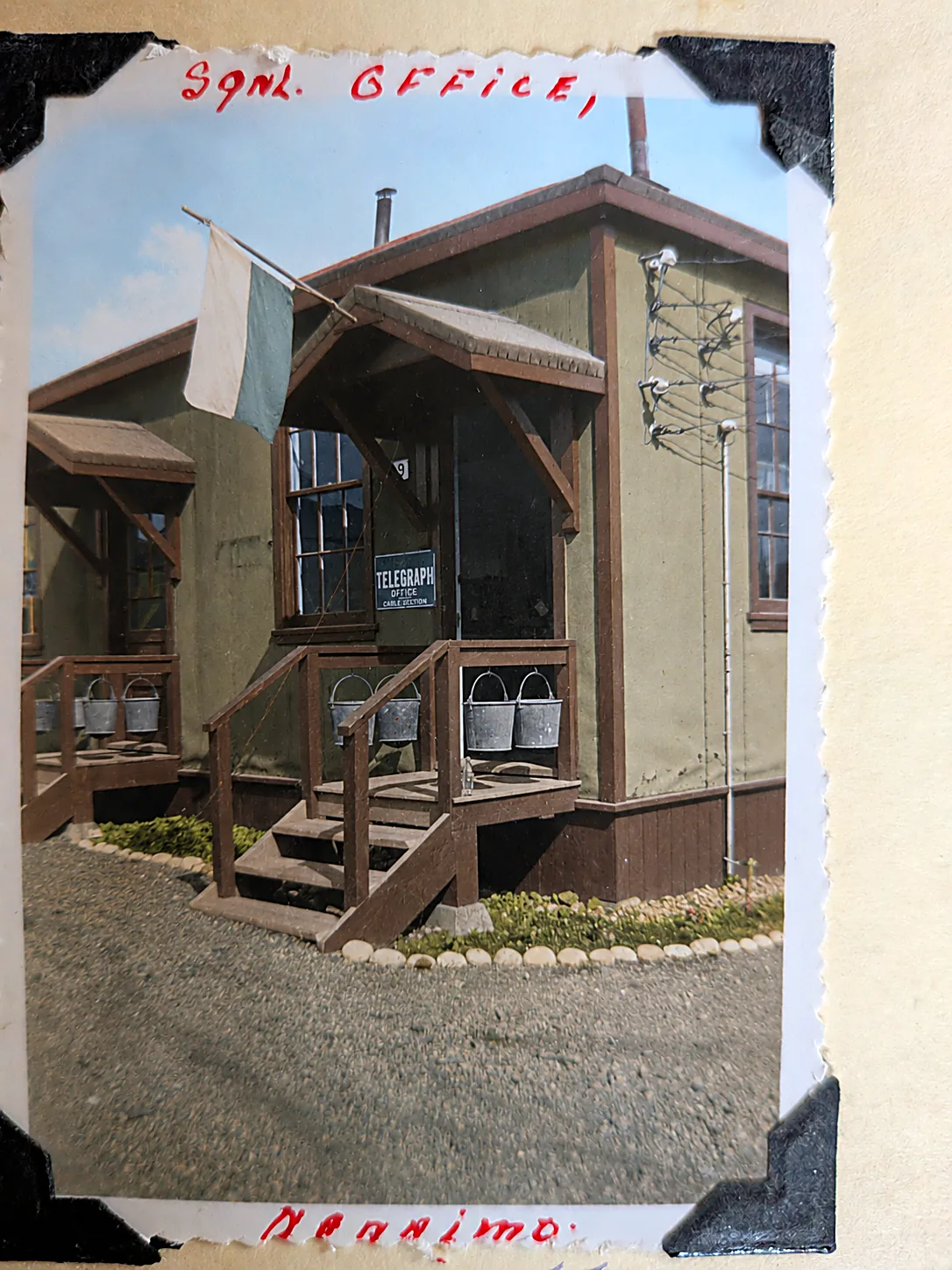 Colourised photograph of a small wooden army building with a flag over the porch and a sign reading Telegraph Office, Cable Section, fire buckets hung along the railing. Captioned Sgnl. Office, Nanaimo.