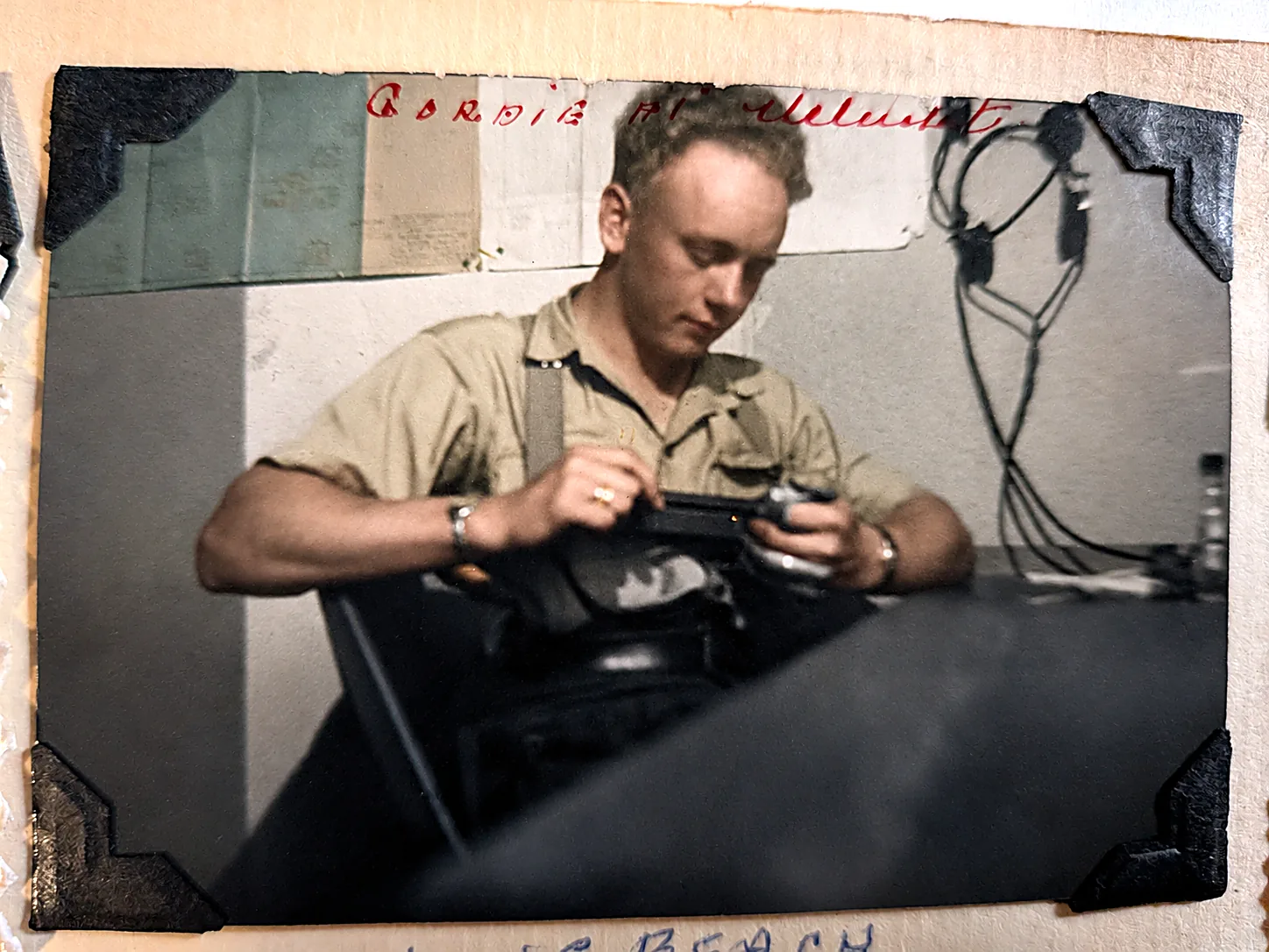 Colourised photograph of Gordie in a light shirt at a desk, working a field telephone or signal set, cables looping on the wall behind him. Long Beach.