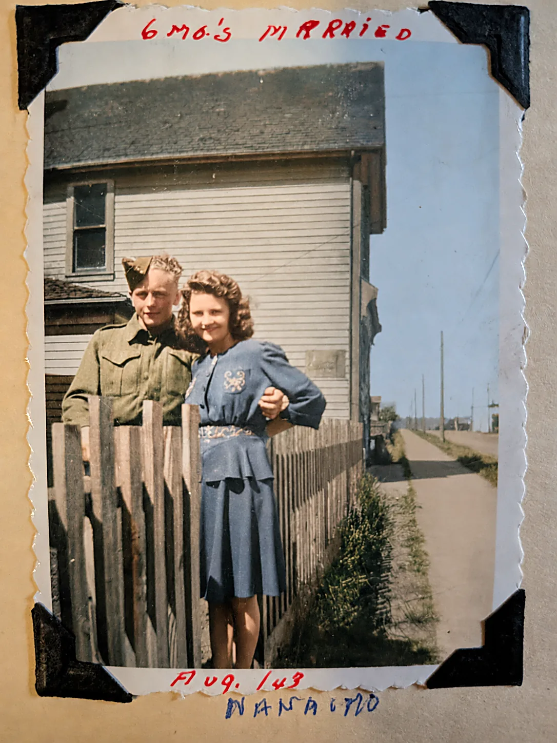 Colourised photograph of Gordie in uniform and Avis in a blue dress standing arm in arm behind a picket fence on a sunny street. Album caption reads 6 mo's married, Aug 1/43, Nanaimo.