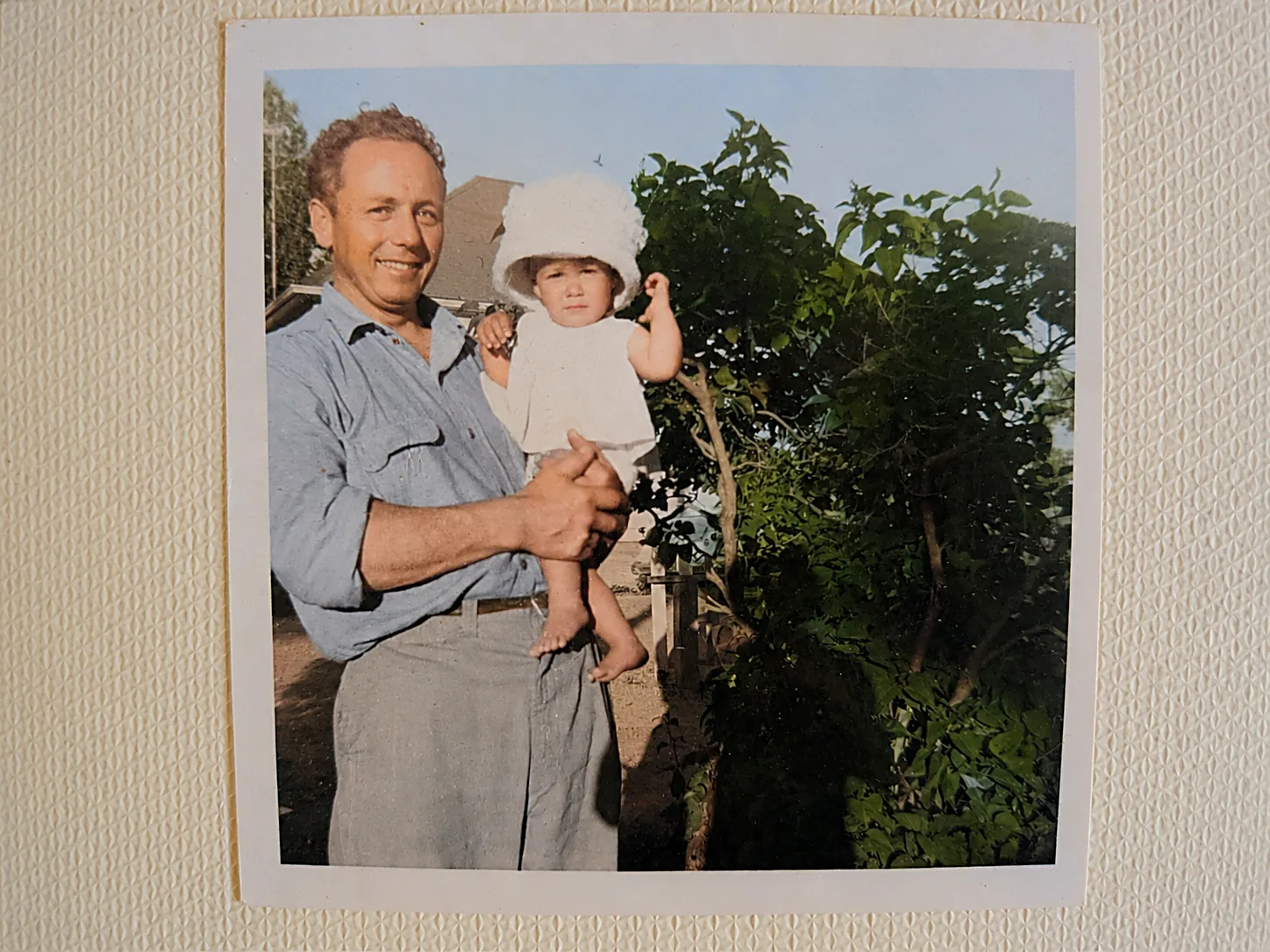 Colourised photograph of Gordie, older and sun-browned in a blue work shirt, holding a toddler in a white sun hat beside a leafy tree in a garden.