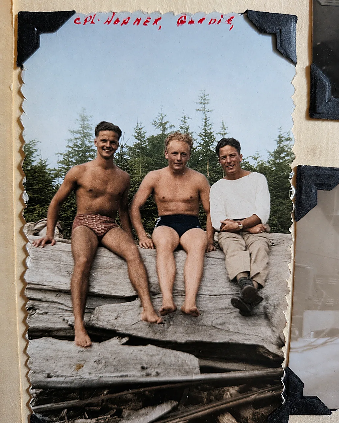 Colourised photograph of three men sitting on bleached driftwood in front of coastal pines, two in swim trunks and one in a white shirt and khakis. Album caption reads Cpl. Horner, Gordie.